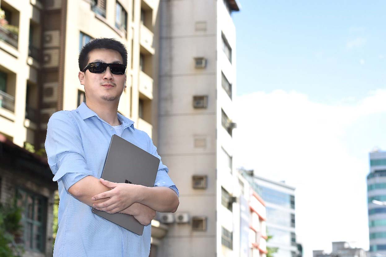 一張含有一位手裡捧者電腦，面露微笑的視障人士的照片