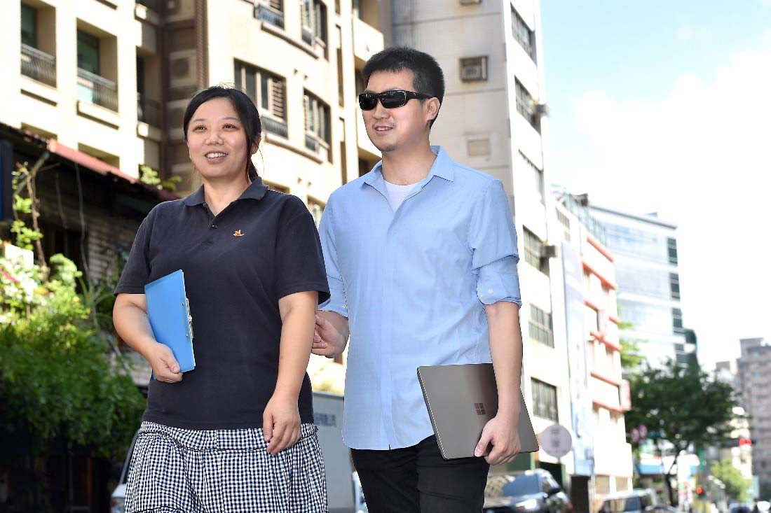 一張含有這位視障人士和一位女士有說有笑並肩走在路上的照片
